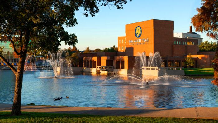 Lake in foreground and brick university building in background with university's lion head logo and the name of the university on the building.